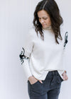 Model wearing a cream sweater with black embroidered flower detail, mock neckline and split sides.