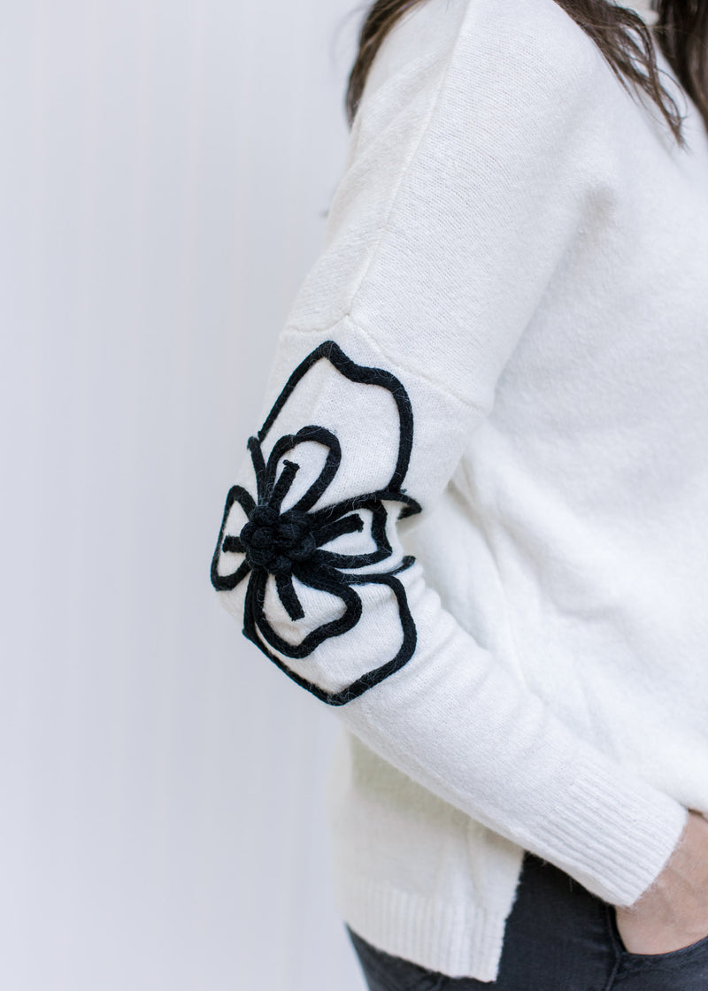 Close up of black embroidered flower on the elbow of a long sleeve cream sweater with split sides. 