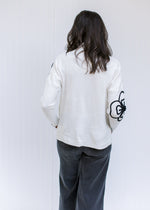 Back view of Model wearing a cream sweater with black embroidered flower detail and a mock neck.
