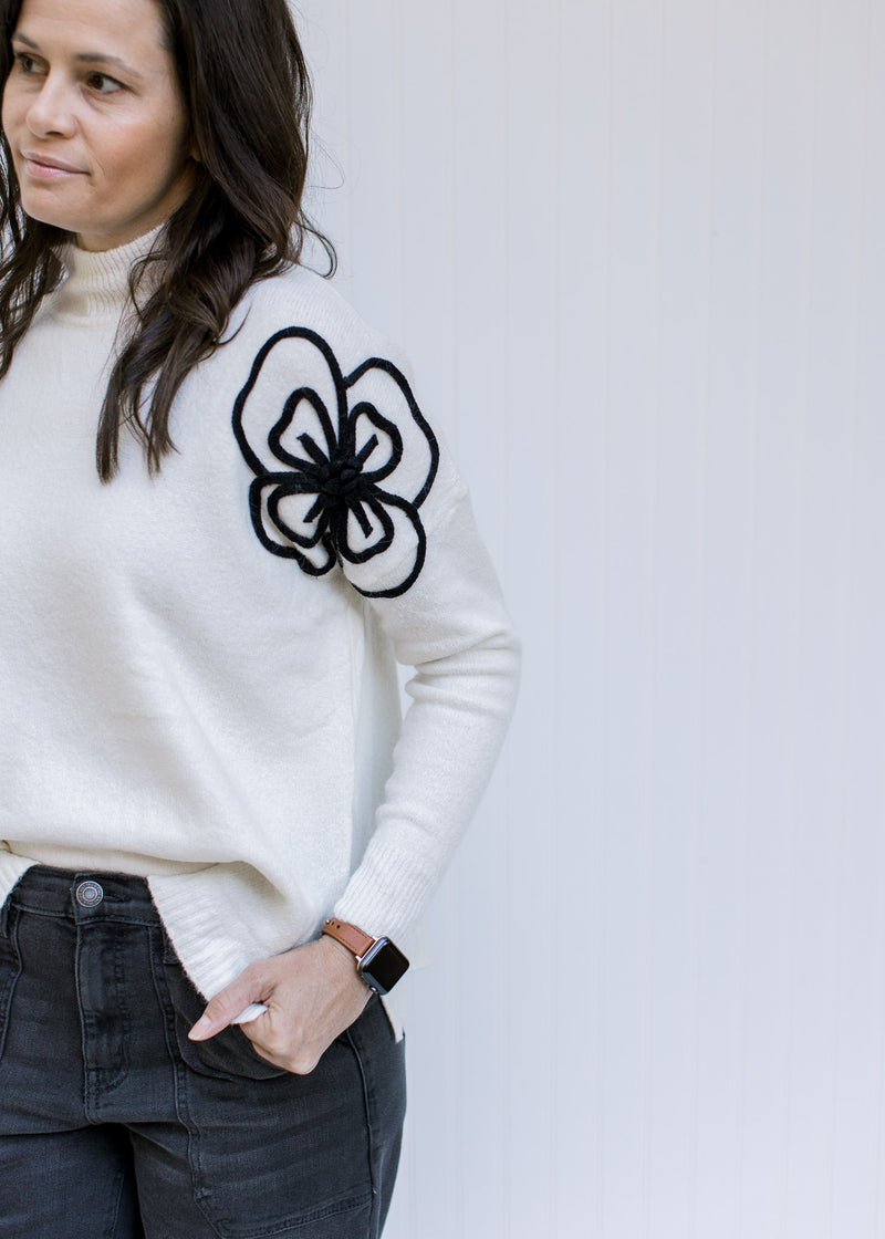 Close up of black embroidered flower on shoulder of a long sleeve cream sweater with split sides.