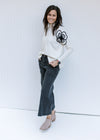 Model wearing jeans and booties with a cream sweater with a black embroidered flower detail. 