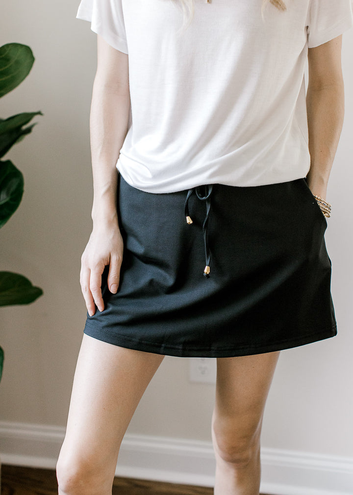 Model wearing a black skort with an elastic waist with tie, built-in shorts and pockets.