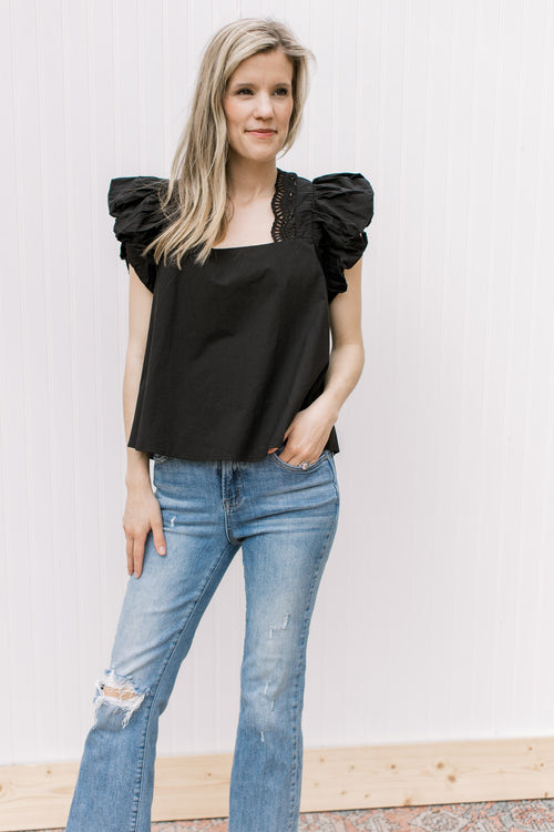 Model wearing a black square neck top with eyelets along the square neck and ruffle cap sleeves.
