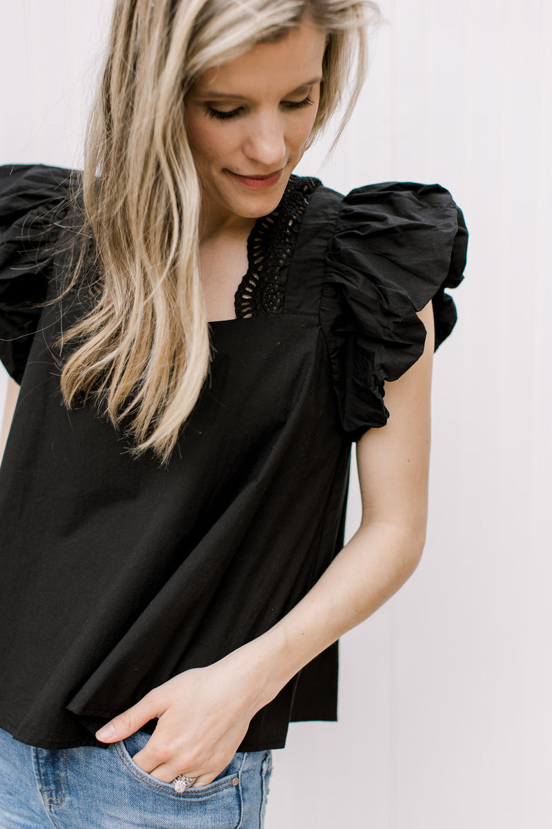 Close up of ruffle cap sleeve and eyelets detail along square neckline of a black top.
