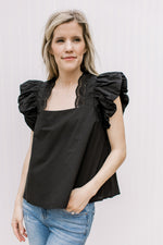 Model wearing jeans with a black top with eyelets along the square neck and ruffle cap sleeves.
