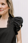 Close up of lace detail along the square neck of a black top with ruffle cap sleeves.