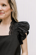 Close up of lace detail along the square neck of a black top with ruffle cap sleeves.