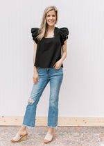 Model wearing jeans, wedges and a black top with eyelets along the neck and ruffle cap sleeves.