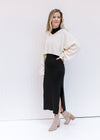 Model wearing a cream sweater with a black sleeveless dress with a mock neckline and a split hem.