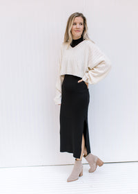 Model wearing a cream sweater with a black sleeveless dress with a mock neckline and a split hem.