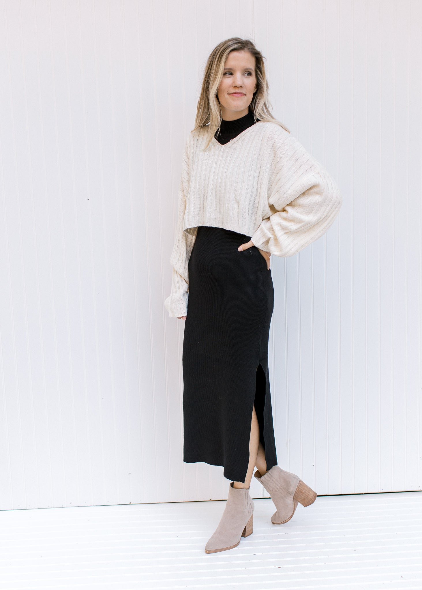Model wearing a cream sweater with a black sleeveless dress with a mock neckline and a split hem.