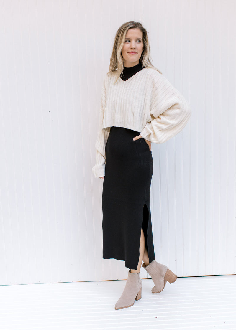 Model wearing a cream sweater with a black sleeveless dress with a mock neckline and a split hem.