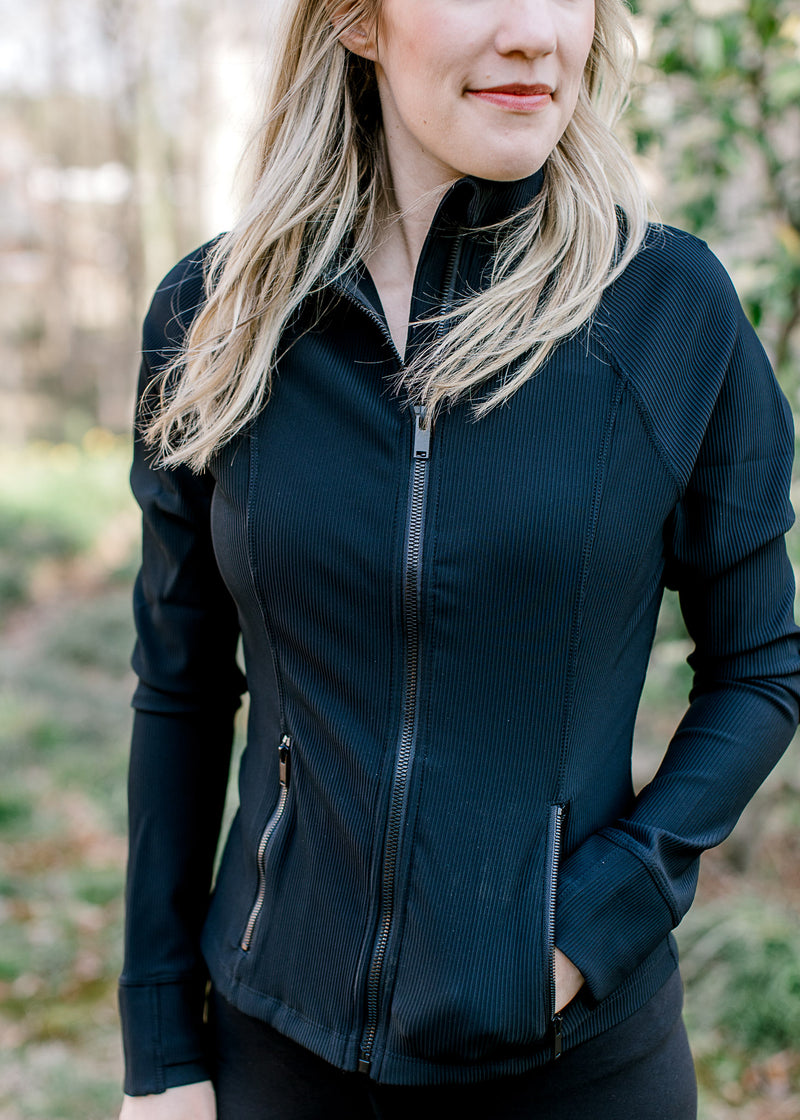 Model wearing a ribbed black jacket with a slim fit, front zip,  zip pockets and long sleeves.