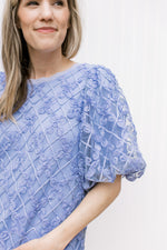 Model wearing a blue top with a sheer floral overlay, bubble short sleeves and a tie back.