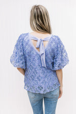 Back view of Model wearing a blue top with a floral overlay, bubble short sleeves and a tie back.