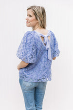 Model wearing jeans with a blue top with a floral overlay, bubble short sleeves and a tie back.