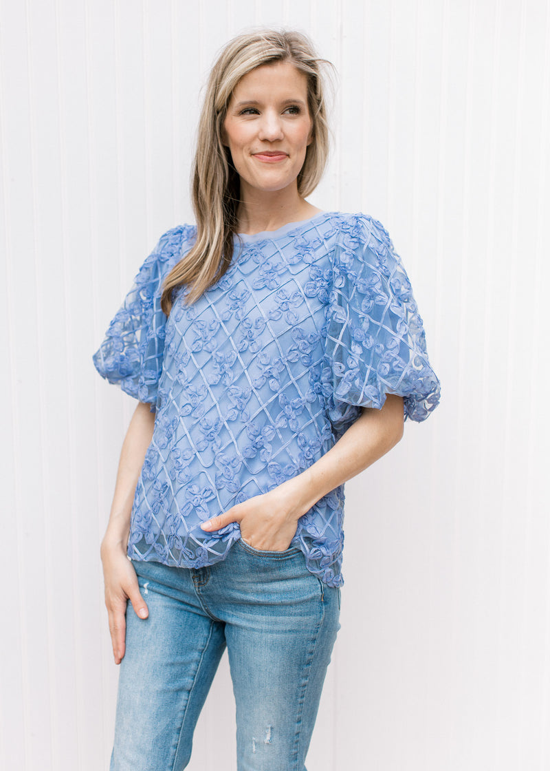 Model wearing a fully lined blue top with a sheer floral overlay and bubble short sleeves.
