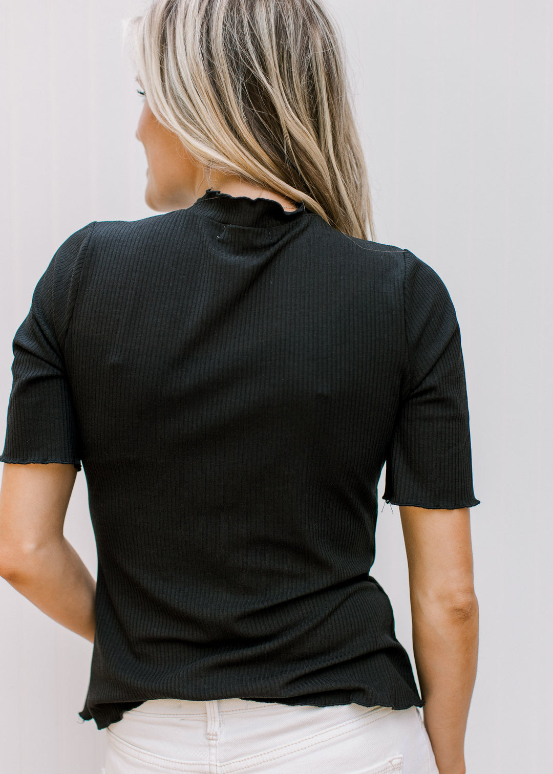 Back view of Model wearing a black ribbed fitted top with short sleeved and ruffle neck and hem.