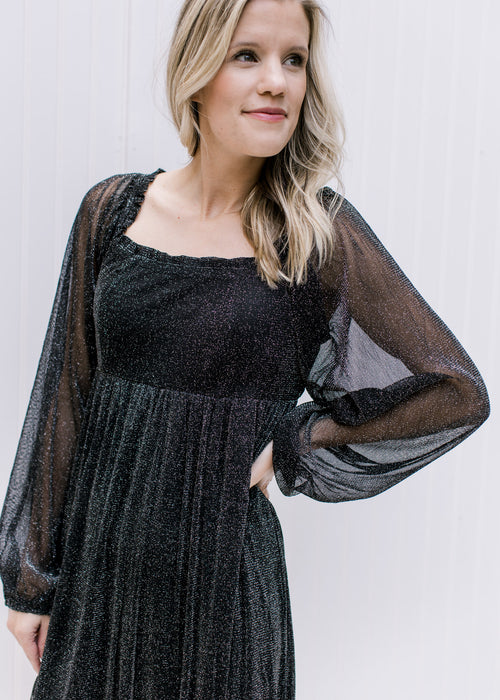 Model in a black shimmery above the knee dress with sheer long sleeves, smocked back with a tie.