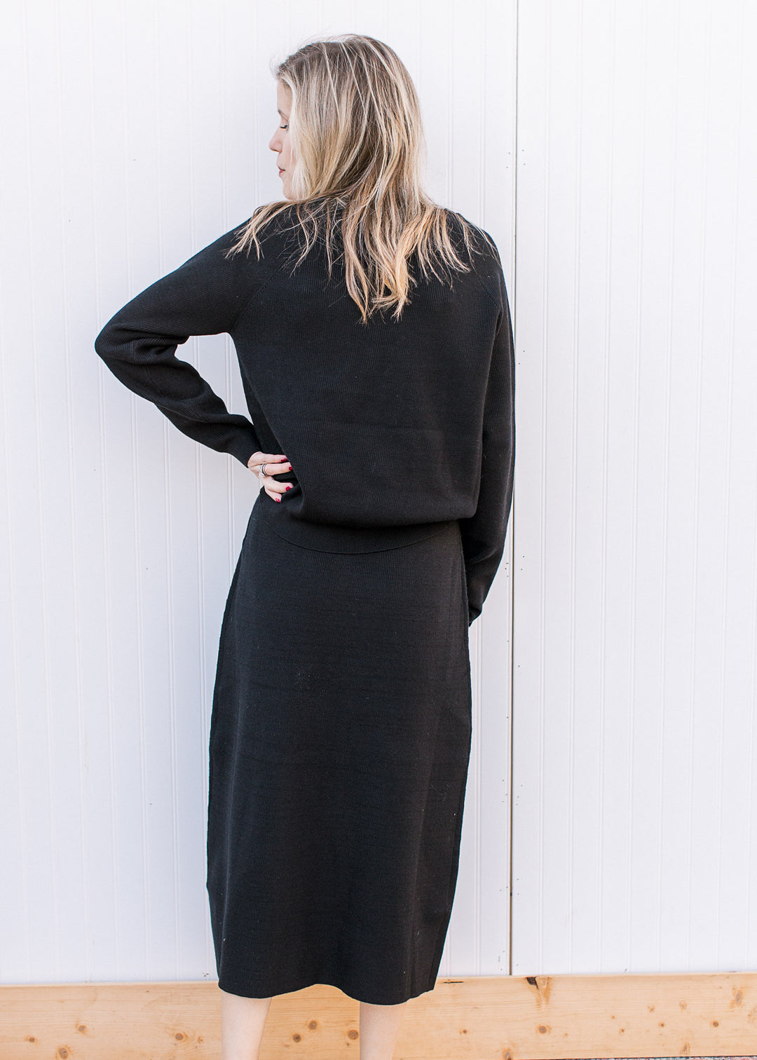 Back view of Model wearing a black ribbed set with midi skirt and long sleeve top with button cuffs.