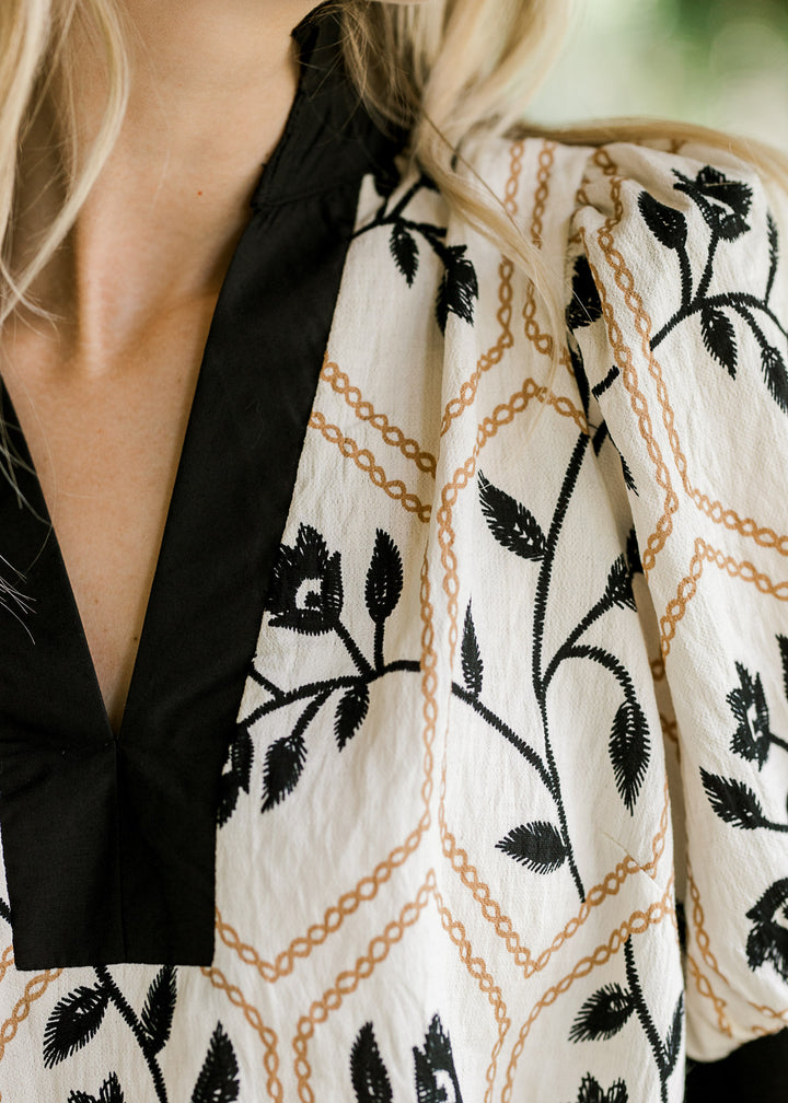 Close up of black trim v-neck with ruffle at neck on a cream top with black floral and gold motif. 