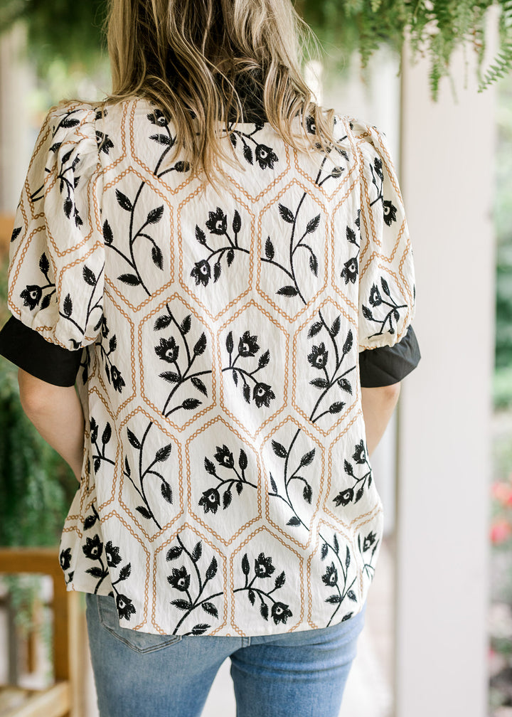 Back view of Model wearing a cream top with black florals inside a gold motif and short sleeves. 
