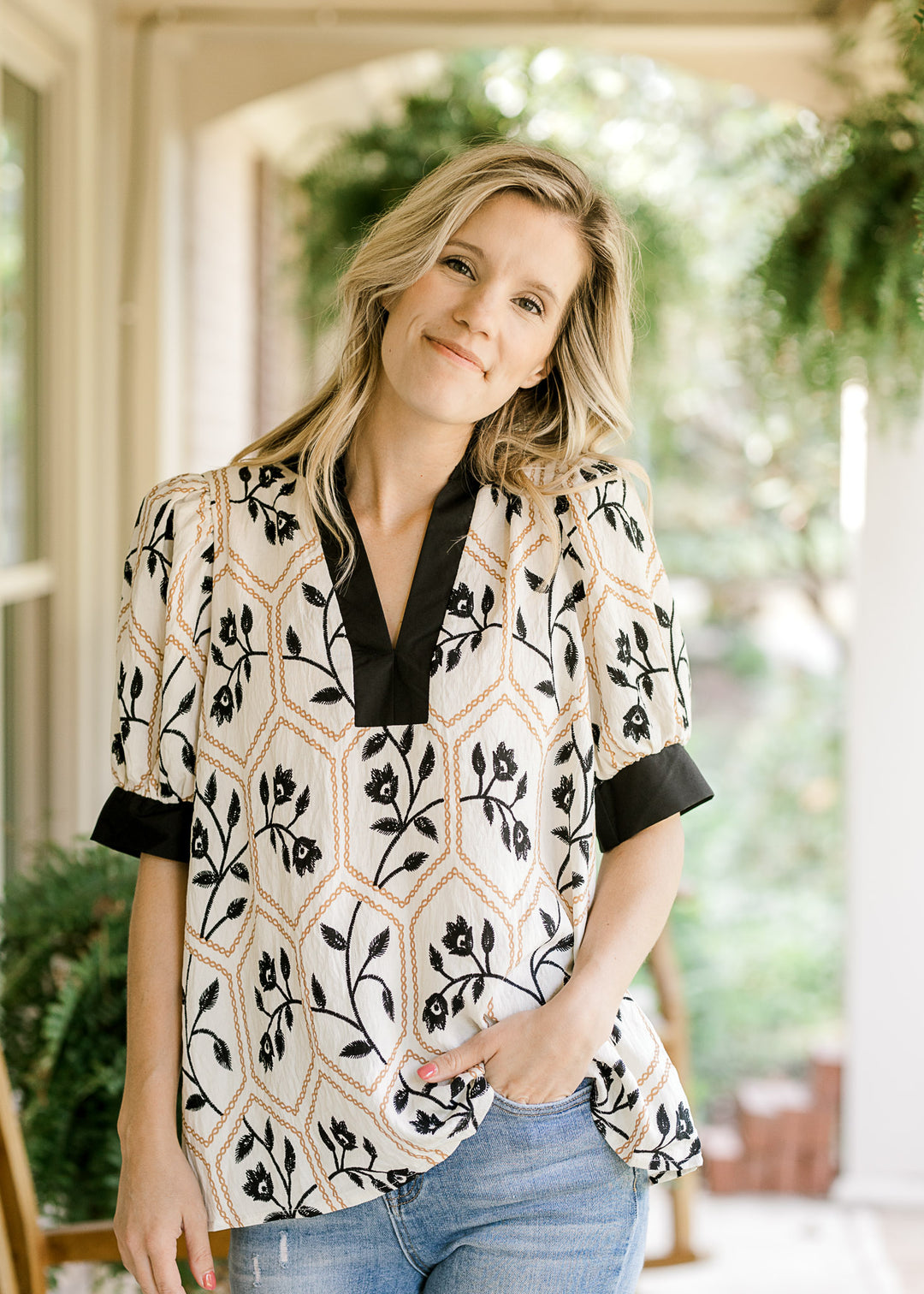 Model wearing a cream v-neck top with black florals inside a gold motif and short sleeves. 