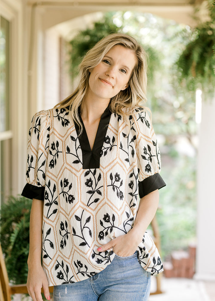 Model wearing a cream v-neck top with black florals inside a gold motif and short sleeves. 