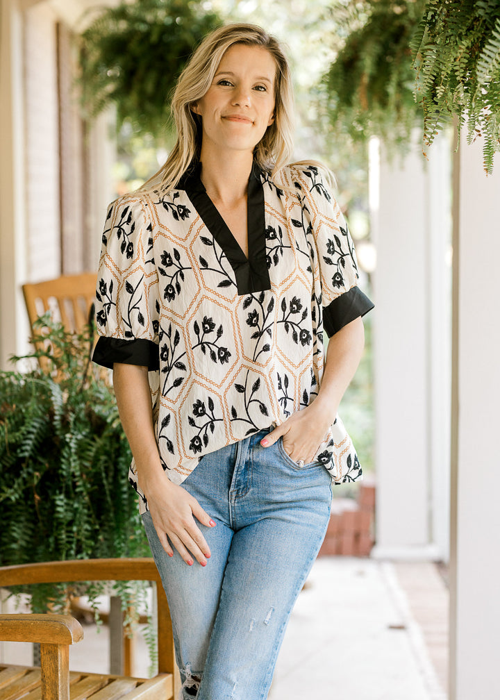 Model wearing a cream top with black florals in gold motif and black trim at neck and cuff. 