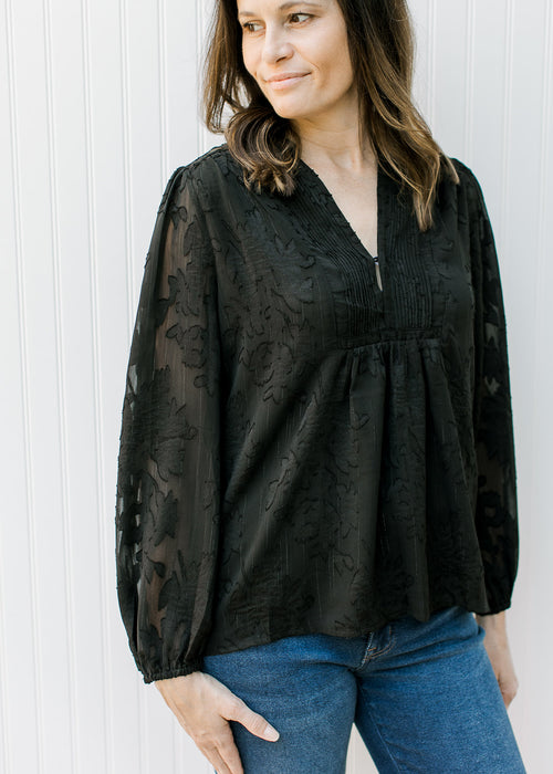 Model wearing a black v-neck top with textured flower pattern and long sheer sleeves. 