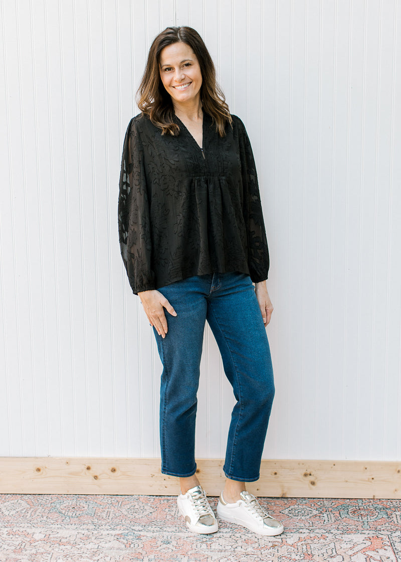 Model wearing jeans and a black v-neck top with textured flower pattern and long sheer sleeves. 