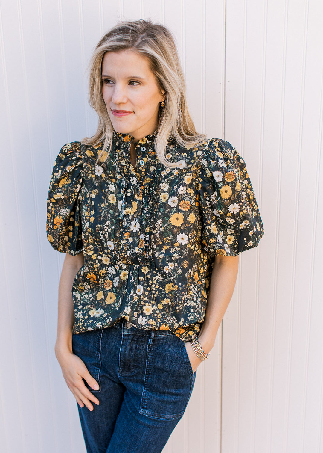Model wearing a black top with yellow and cream floral, buttons, short sleeves and ruffle mock neck.