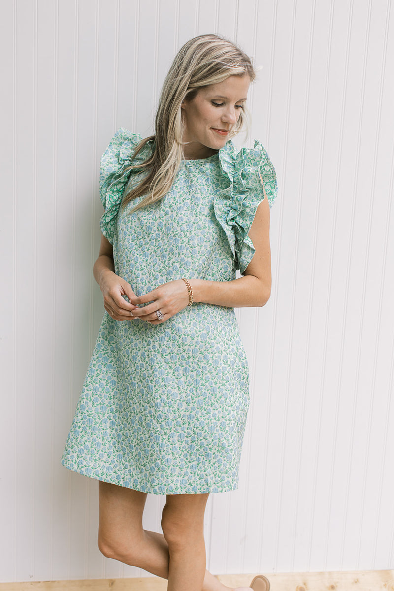 Model wearing a bright green above the knee dress dress with blue floral and sleeveless ruffles. 