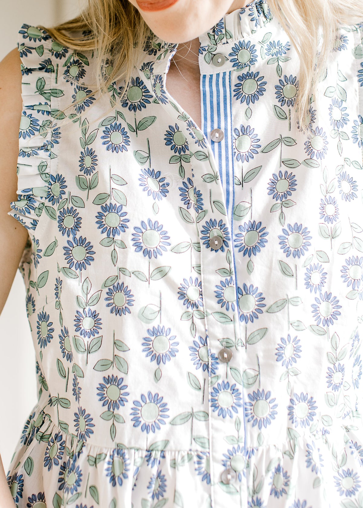 Close up of buttons and ruffles at arm, neck and cuff of a sleeveless white top with flowers. 