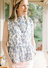 Model wearing a sleeveless white top with blue and green sunflowers, ruffle detail and buttons. 