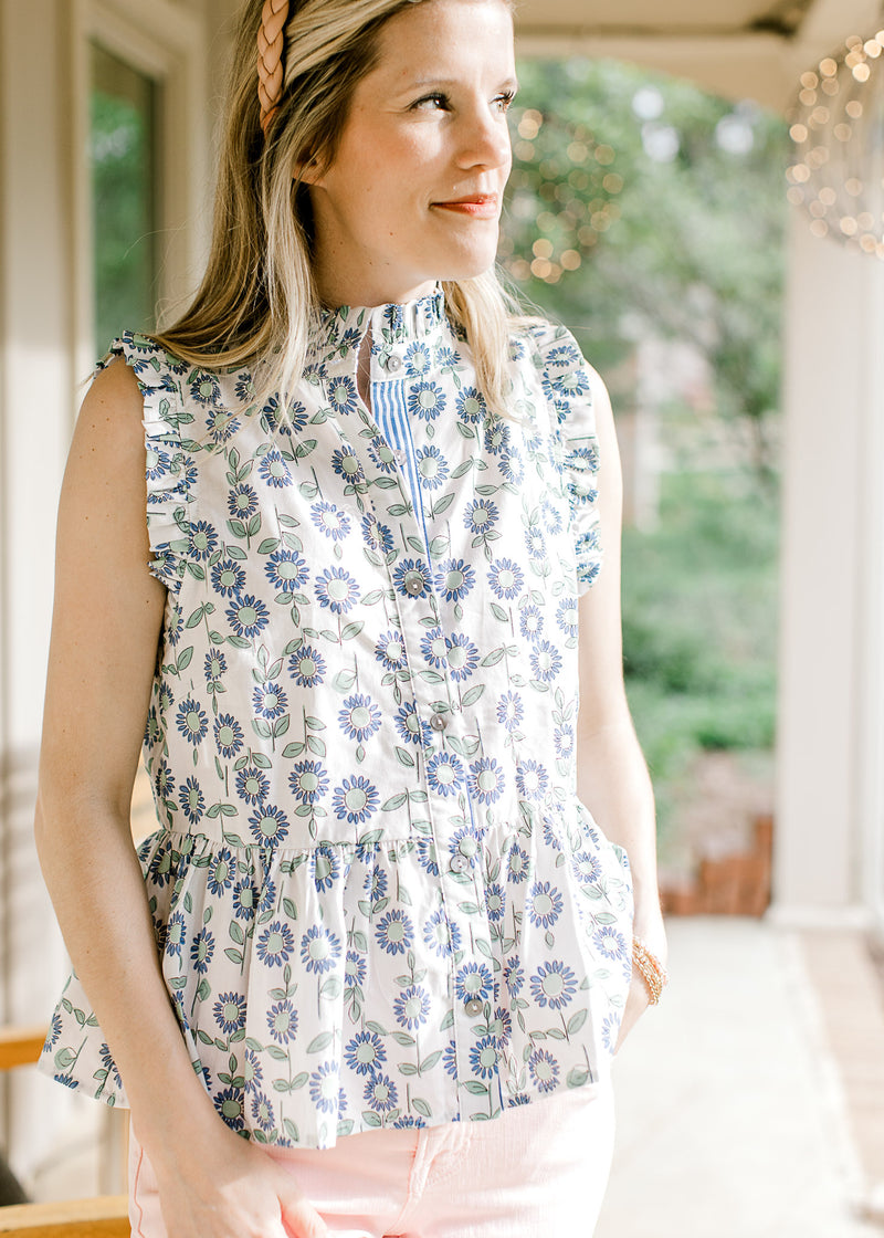 Model wearing a sleeveless white top with blue and green sunflowers, ruffle detail and buttons. 