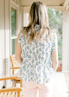 Back view of Model wearing a white top with blue and green sunflowers, ruffle detail and buttons. 