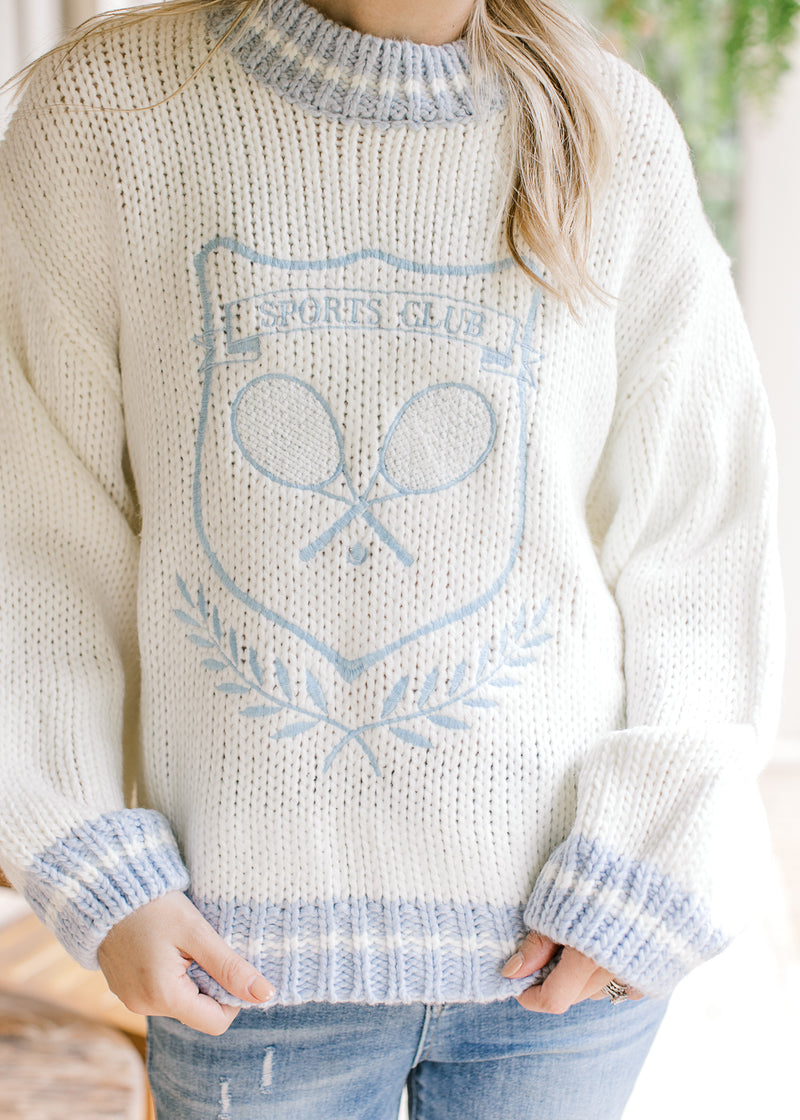 Model wearing a white sweater with pale blue embroidered tennis design and long sleeves. 