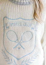 Close up of embroidered pale blue tennis design on a white knit long sleeve sweater.