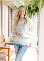 Model wearing jeans with a white sweater with pale blue embroidered tennis design. 