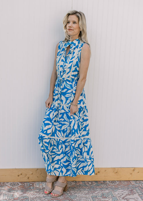 Model wearing a sleeveless cream and blue floral maxi with lace inset at bodice and tie waist.
