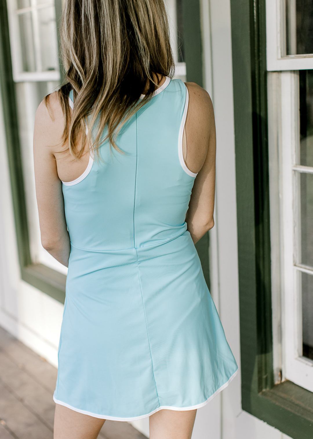 Back view of Model wearing a blue, sleeveless dress with white piping detail and hidden zipper. 