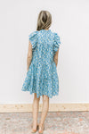 Back view of Model wearing a mint green dress with blue floral, mock neck and ruffle cap sleeves.