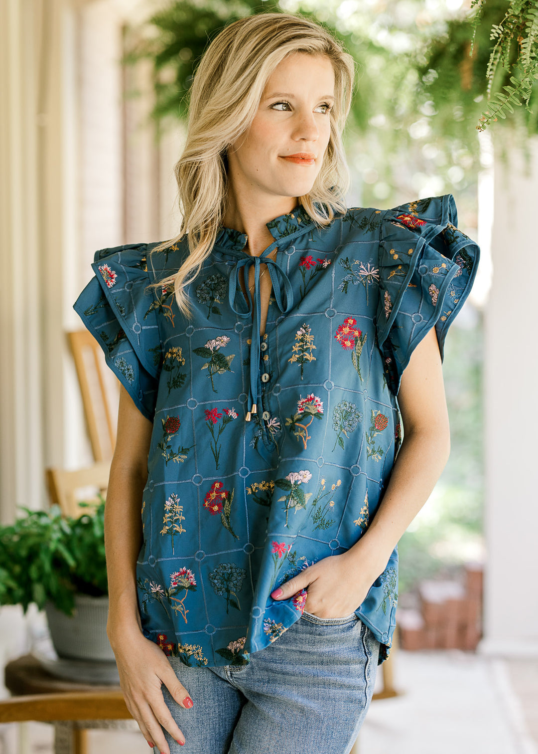 Model wearing a blue top with embroidered squares with floral and v-neck with a tie. 
