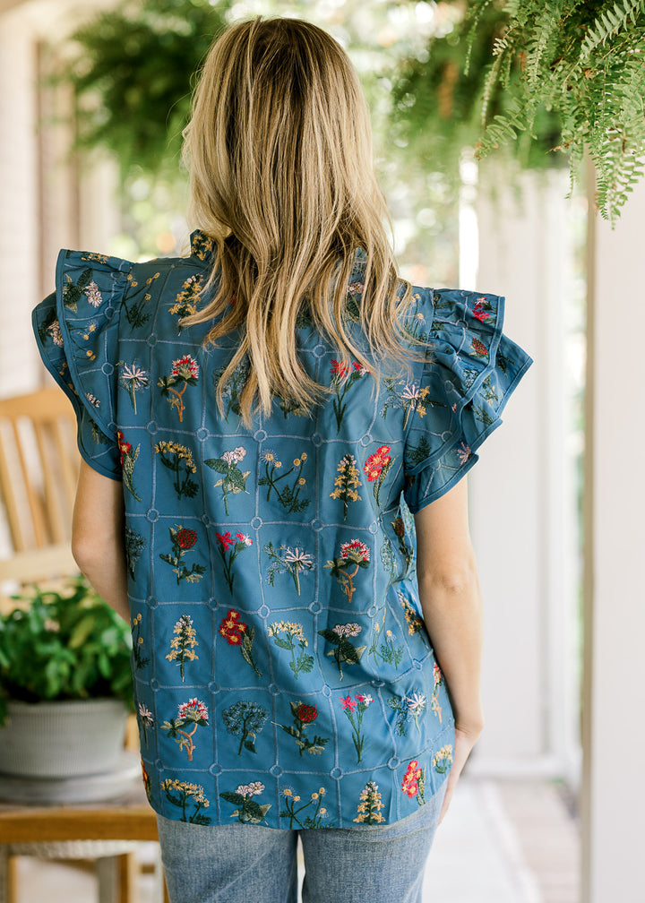 Back view of Model wearing a blue top with embroidered  floral, cap sleeves and v-neck with a tie. 