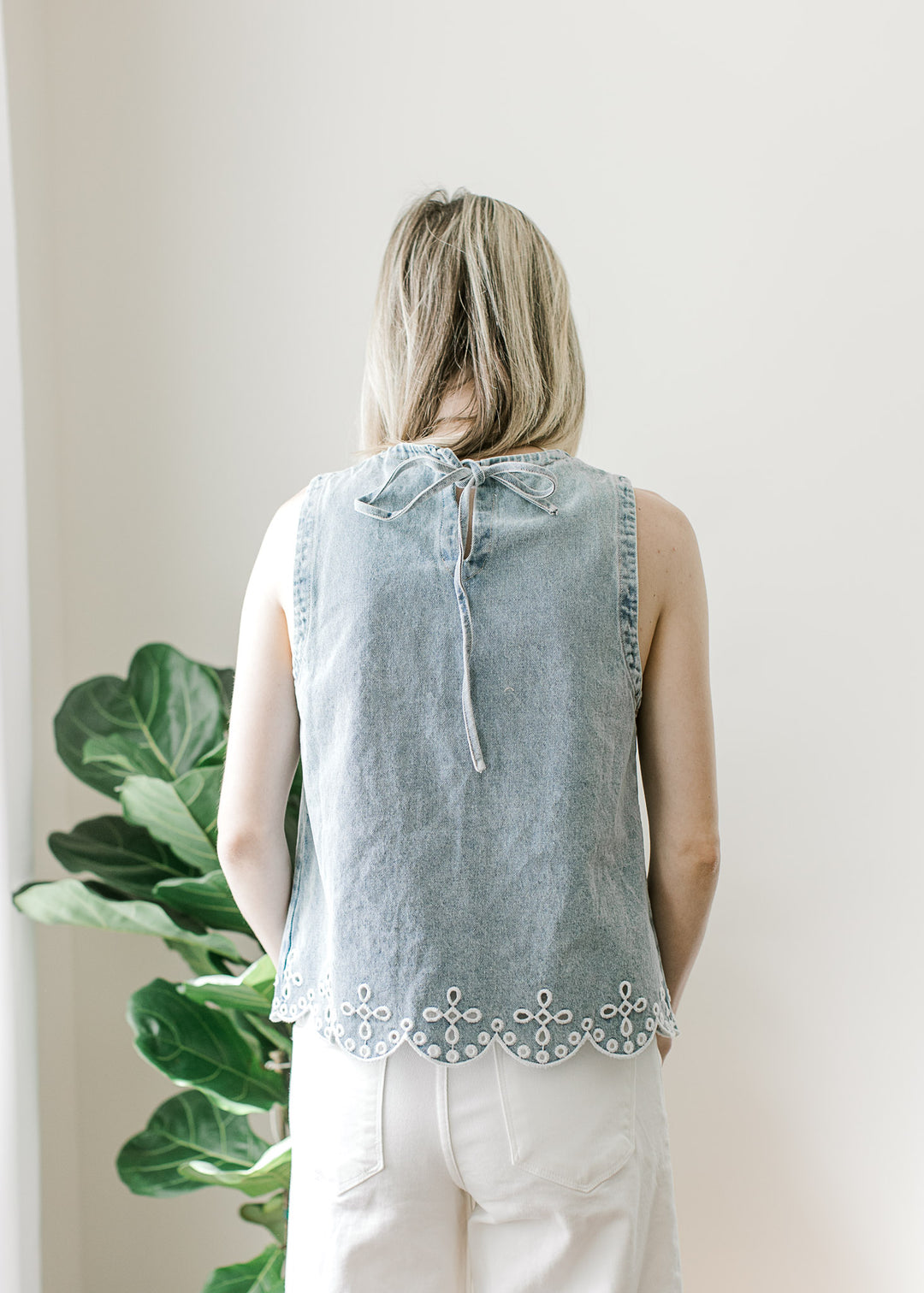 Back view of Model wearing a light wash sleeveless top with scalloped hem and tie keyhole. 