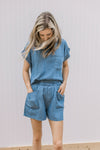 Model wearing a chambray set with patch pockets on the shorts and top and rolled short sleeves.