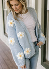 Model wearing a white top with a knit cornflower blue cardigan with white daisies. 