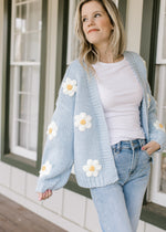 Model wearing jeans a knit cornflower blue cardigan with white daisies and long sleeves. 