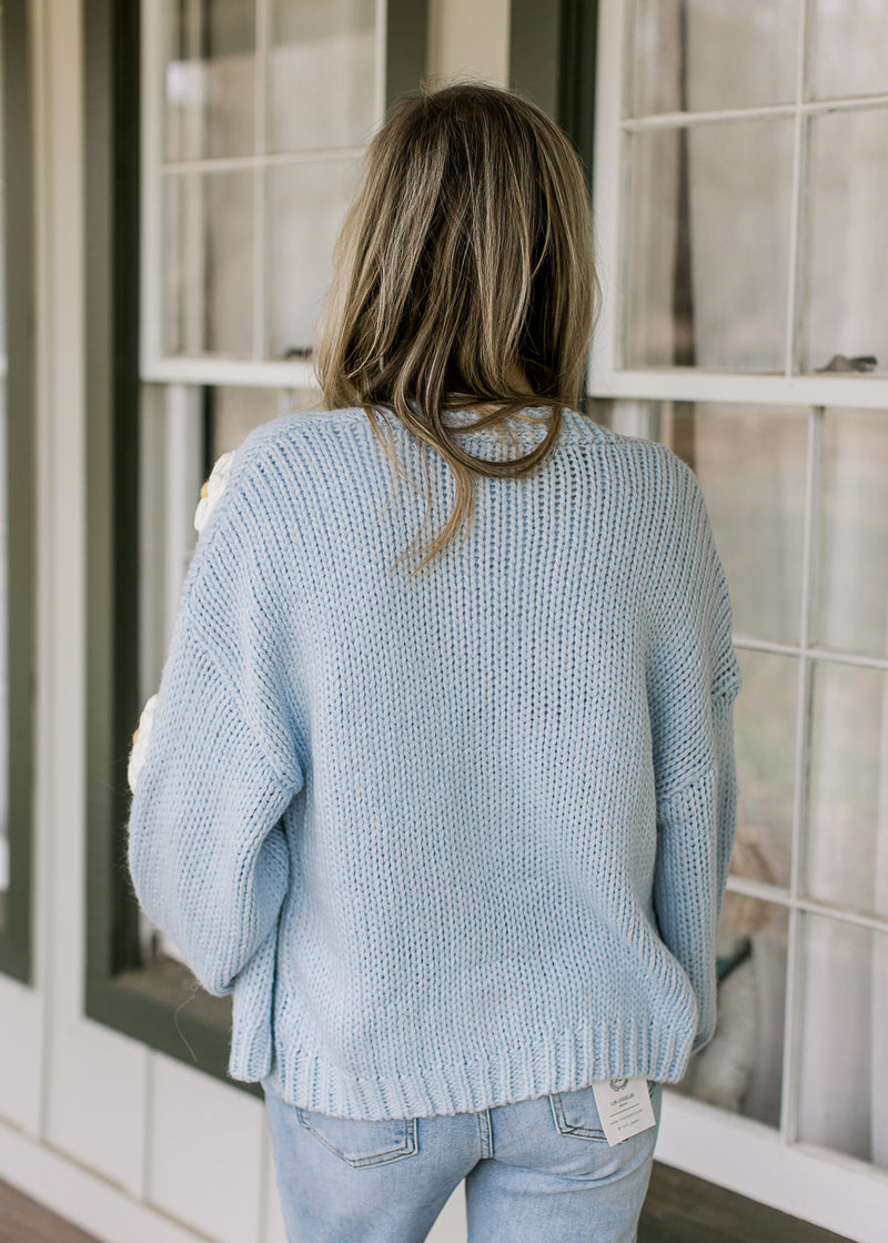 Back view of Model wearing a knit pale blue cardigan with white daisies and long sleeves. 